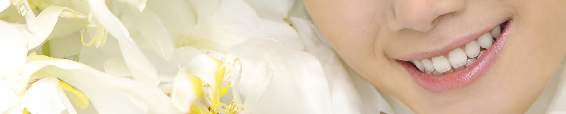 A woman with bright white teeth smiling at the camera, set against a blurred background featuring white flowers.