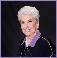 The image displays a woman with short hair, wearing a dark jacket over a purple top, smiling at the camera against a black background. She has a white necklace and appears to be middle-aged.