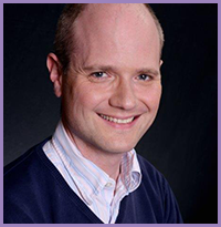 The image shows a man smiling at the camera, with a bald head, wearing a dark blue shirt, and standing against a black background.