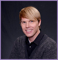 The image shows a man with blond hair smiling at the camera. He has a beard and mustache, and he's wearing a dark top with a collar. There is no text present in the image.