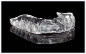 The image shows a clear acrylic dental retainer with multiple teeth-shaped openings, placed on a black background.