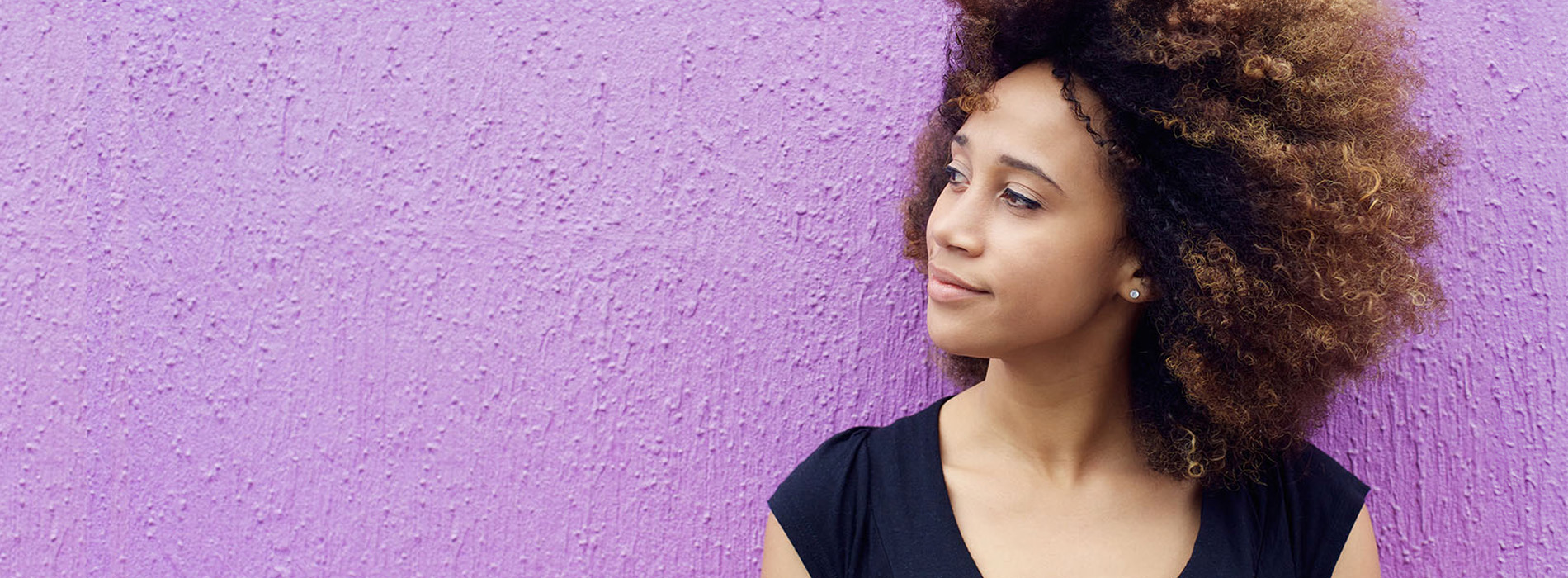 A woman with curly hair stands against a purple wall, captured from behind.