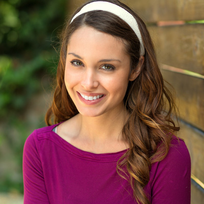 The image shows a woman with long hair smiling at the camera, wearing a purple top and standing against a wooden fence.