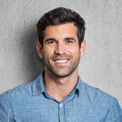 A young man with facial hair, smiling broadly, poses against a grey wall with a blue denim shirt.