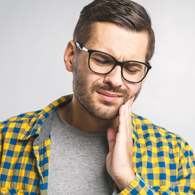 This image features a man with glasses, wearing a yellow plaid shirt, holding his hand to his chin in a contemplative pose, with a concerned expression on his face.