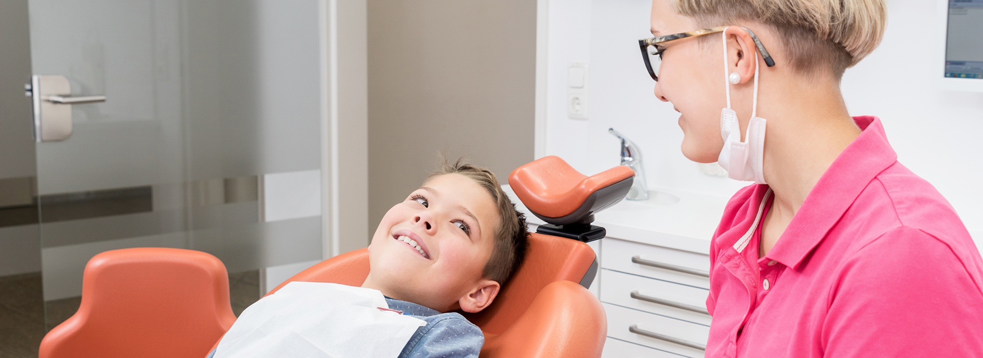 A young boy sitting in a dentist's chair receiving dental care from a woman wearing a pink shirt.