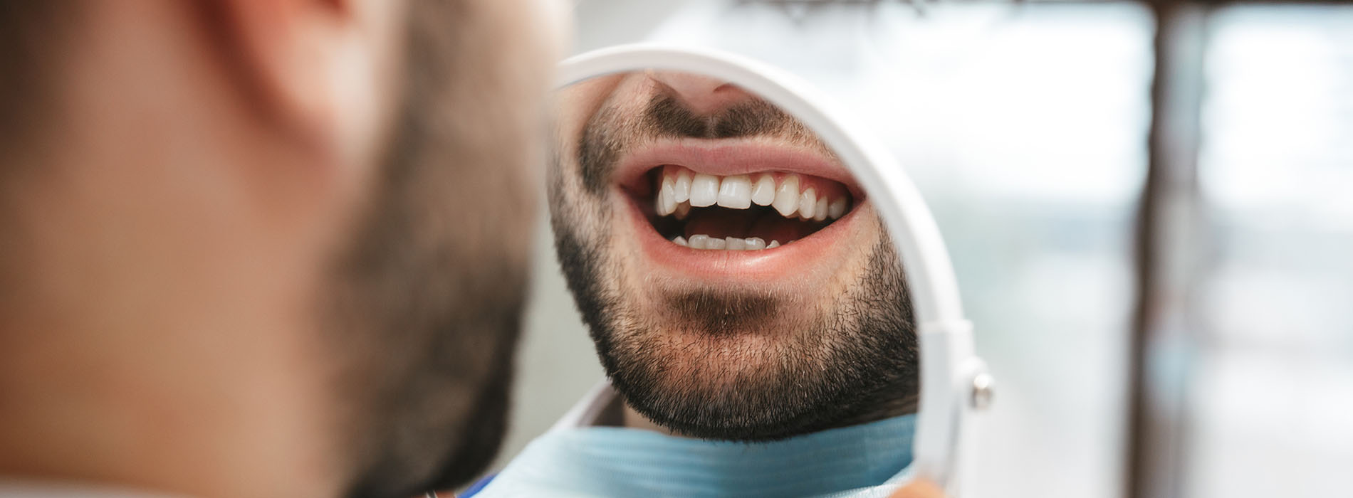 A man with a toothy smile is seen through a mirror reflection, holding a toothbrush, with another person looking at him from behind.