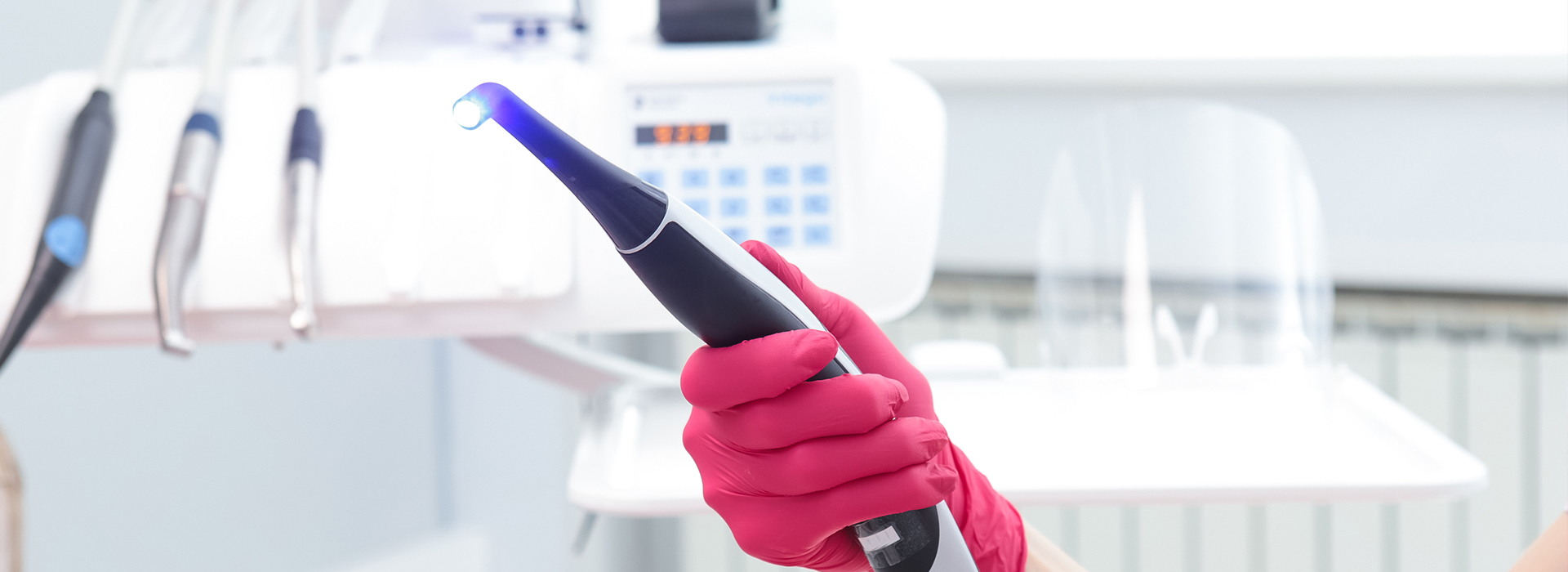 The image shows a person wearing pink gloves holding a black device with a blue light, standing in front of a white countertop with medical equipment on it.