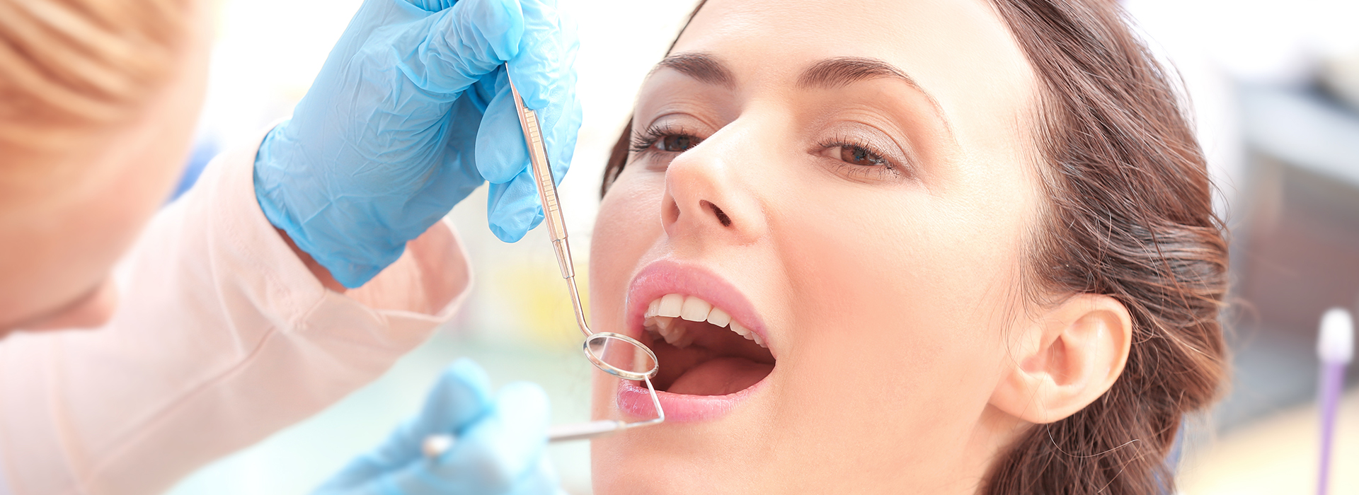 A dental hygienist cleaning a woman's teeth with a dental pick while she sits in a dental chair.