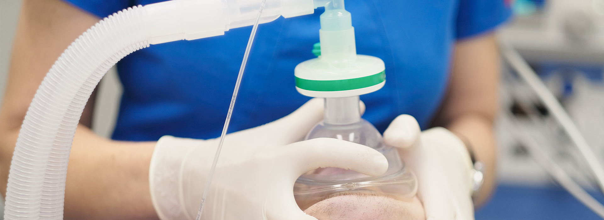 A person wearing gloves and a blue shirt is holding a clear plastic bag with a green cap containing a white substance, which appears to be a medical product, possibly a medication or solution, against a backdrop that includes a machine with tubes and a blue wall.